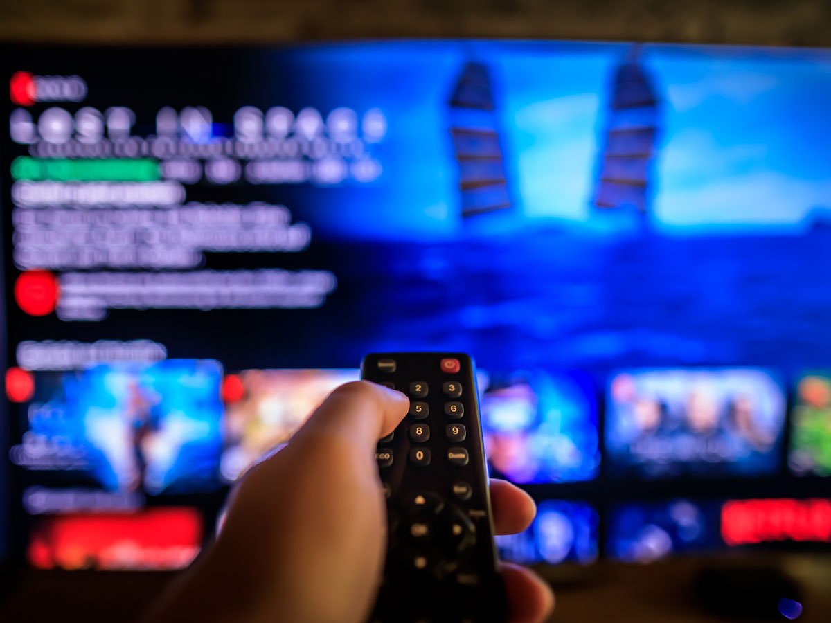 Hand hält Fernbedienung vor unscharfem Fernseher mit Streaming-Inhaltsauswahl.
