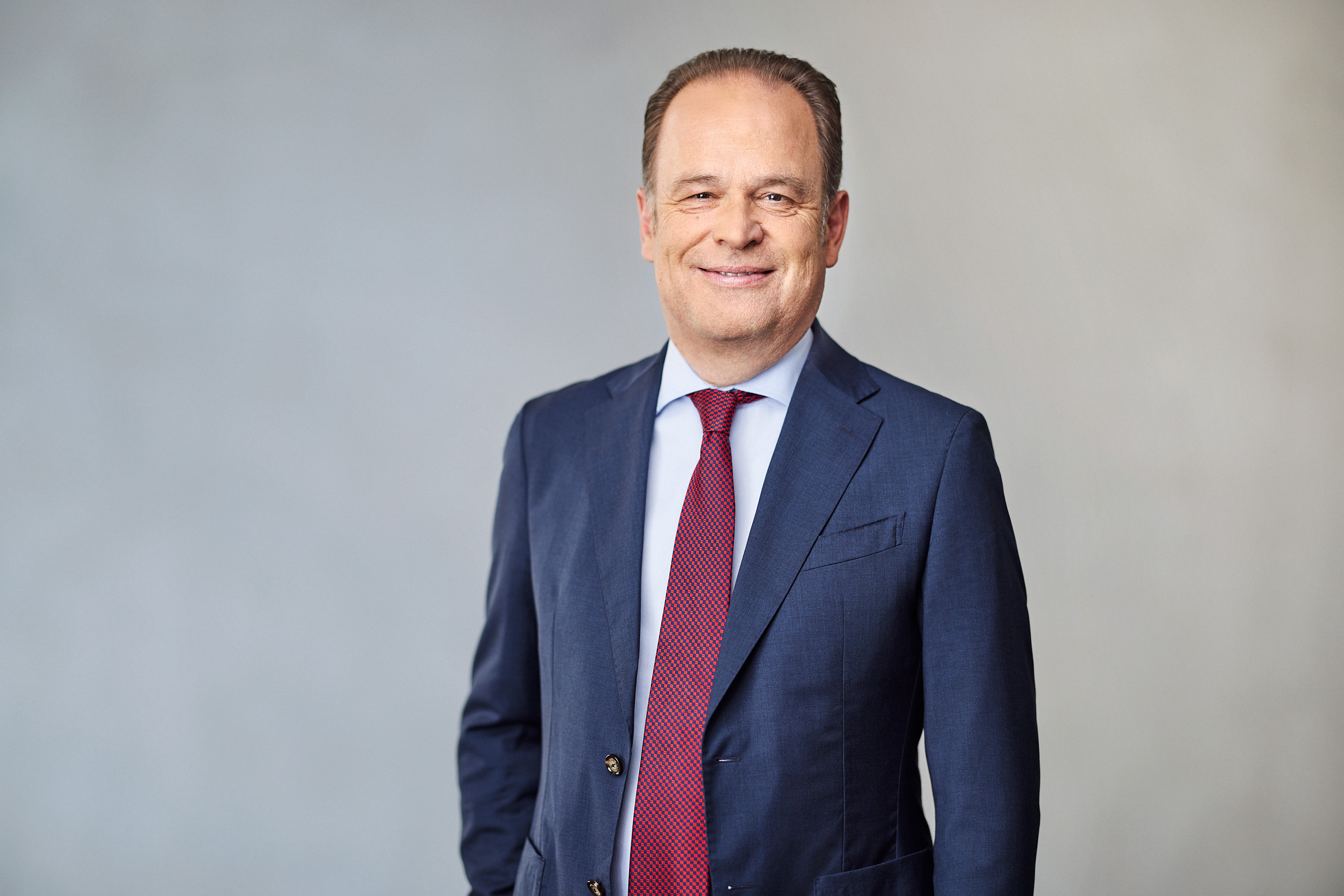 Ein Mann im blauen Anzug mit roter Krawatte lächelt vor neutralem Hintergrund.