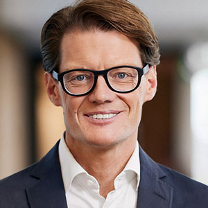 Eine Person mit Brille, die einen Anzug und ein weißes Hemd trägt, lächelt in einer unscharfen Innenumgebung.