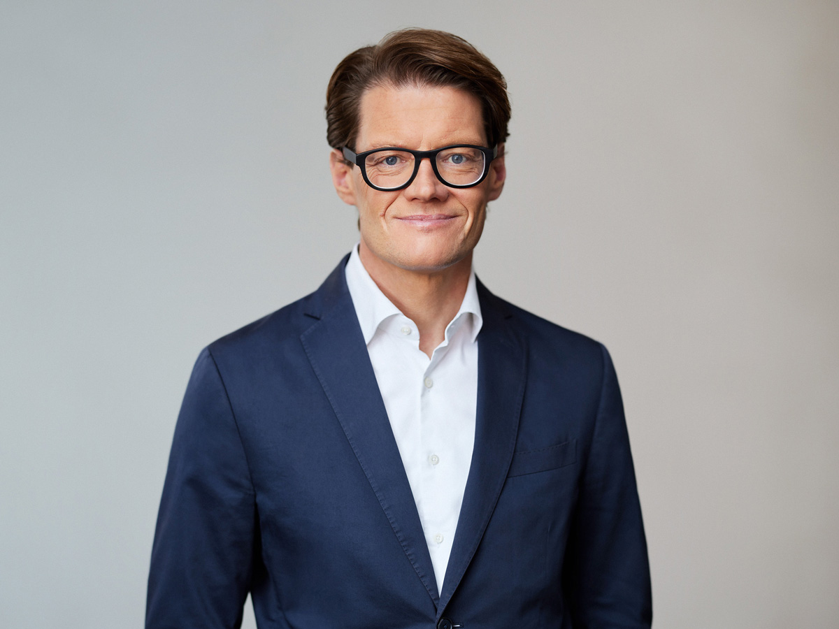 A person in a navy suit and glasses smiles against a plain background.