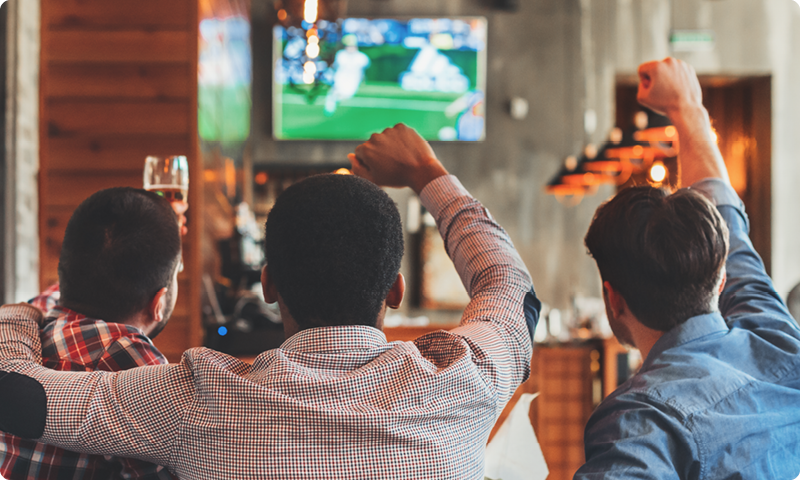 Drei Männer jubeln beim Fußballschauen in einer Bar, ein Mann hält ein Glas.