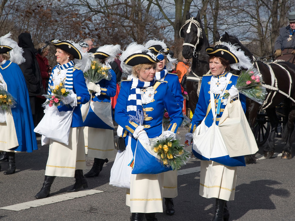Personen in blauen Uniformen mit Blumensträußen marschieren bei einer Parade, Pferdekutsche im Hintergrund.