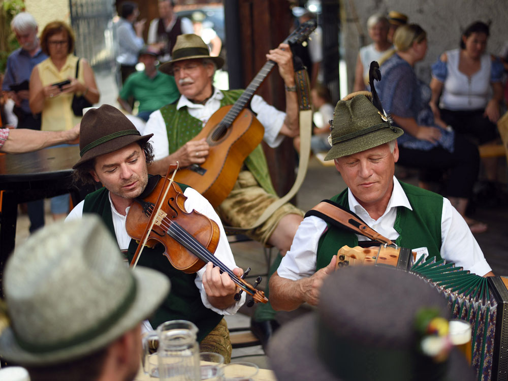 Musiker in Tracht spielen Geige und Akkordeon, im Hintergrund sitzt ein Gitarrist, umgeben von Publikum.