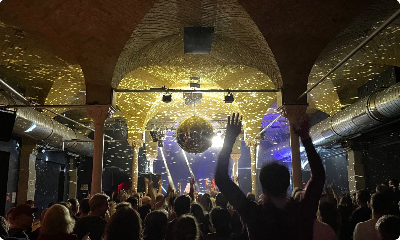 Menschen tanzen in einem Club mit Discokugel und Lichteffekten unter gewölbten Decken.