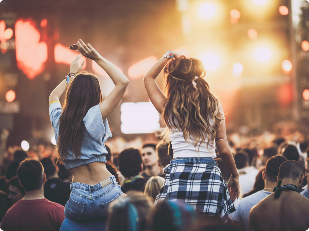 Zwei junge Frauen von hinten auf den Schultern anderer Menschen bei einem Konzert mit hellen Bühnenlichtern im Hintergrund.