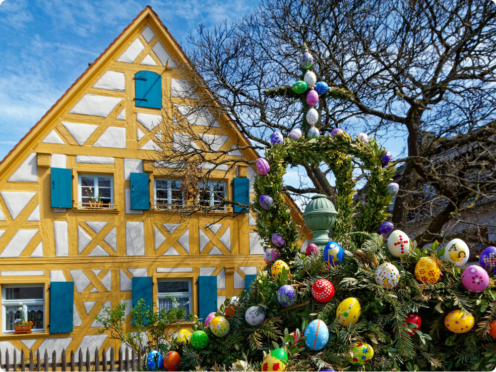 Osterbrunnen mit bunten Ostereiern und grünen Zweigen vor einem Fachwerkhaus mit gelben Balken und blauen Fensterläden an einem sonnigen Frühlingstag.