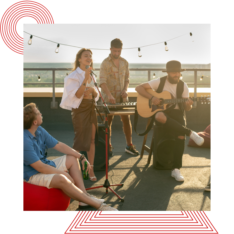 Gruppe junger Menschen auf einer Dachterrasse mit einer Frau am Mikrofon, einem Mann an einem Keyboard und einem weiteren Mann mit Gitarre, umgeben von Sitzgelegenheiten und Lichterkette.
