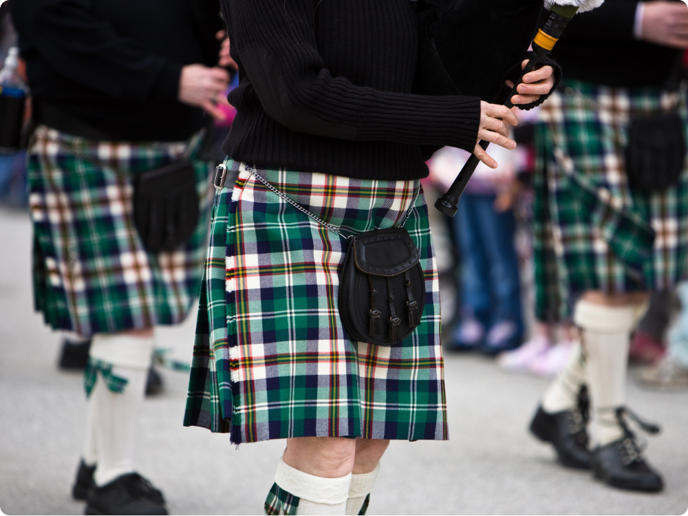 Person in schwarzem Pullover und grün-kariertem Kilt mit schwarzer Tasche spielt Dudelsack.