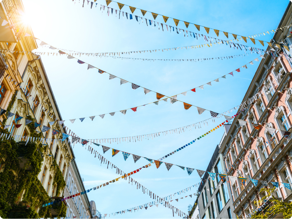Mehrere Reihen bunter Wimpel hängen quer zwischen zwei Häuserzeilen unter blauem Himmel