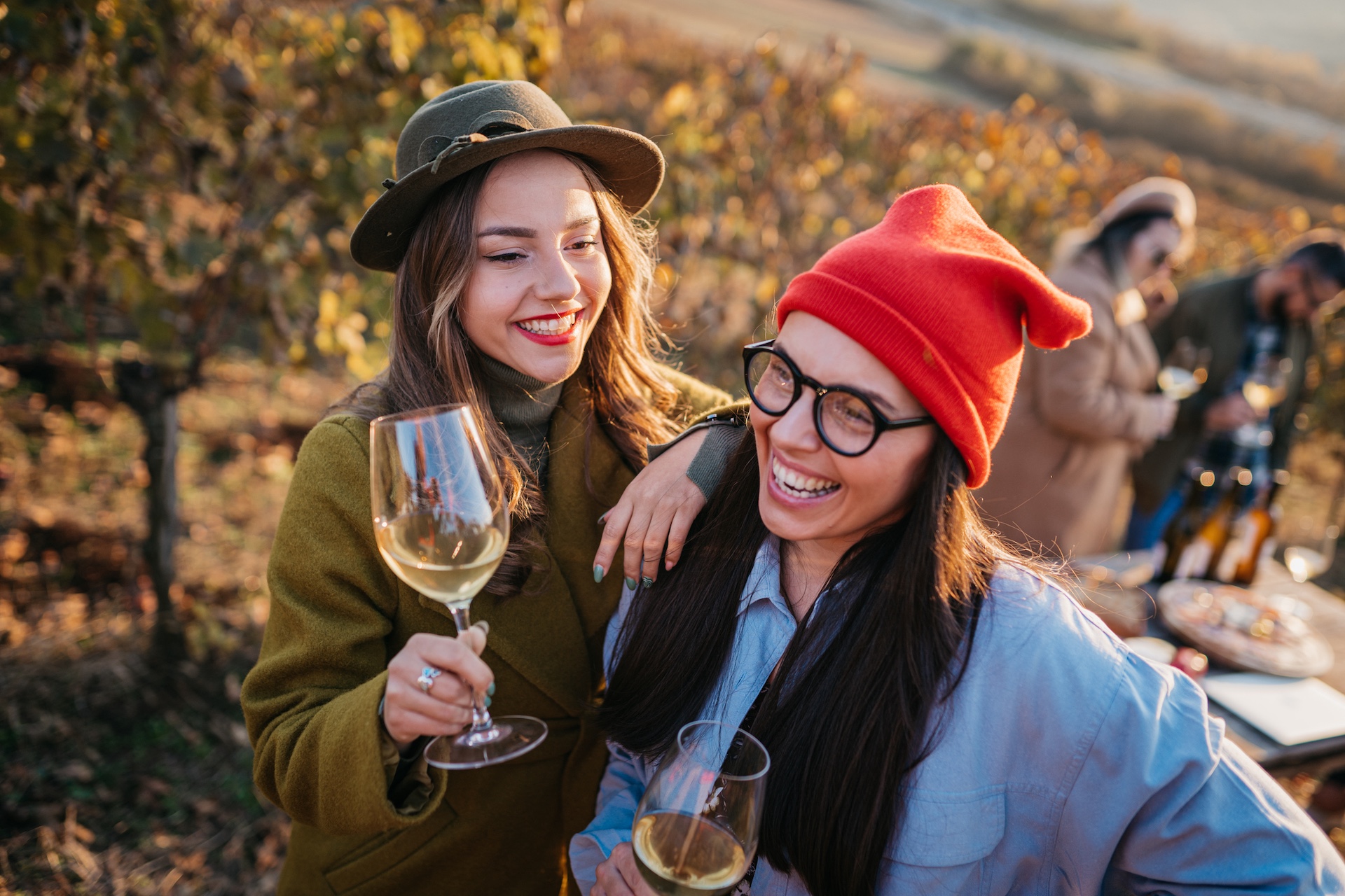 Zwei Frauen mit Wein in der Hand lachen in einem Weinberg, im Hintergrund zwei weitere Personen mit Wein.