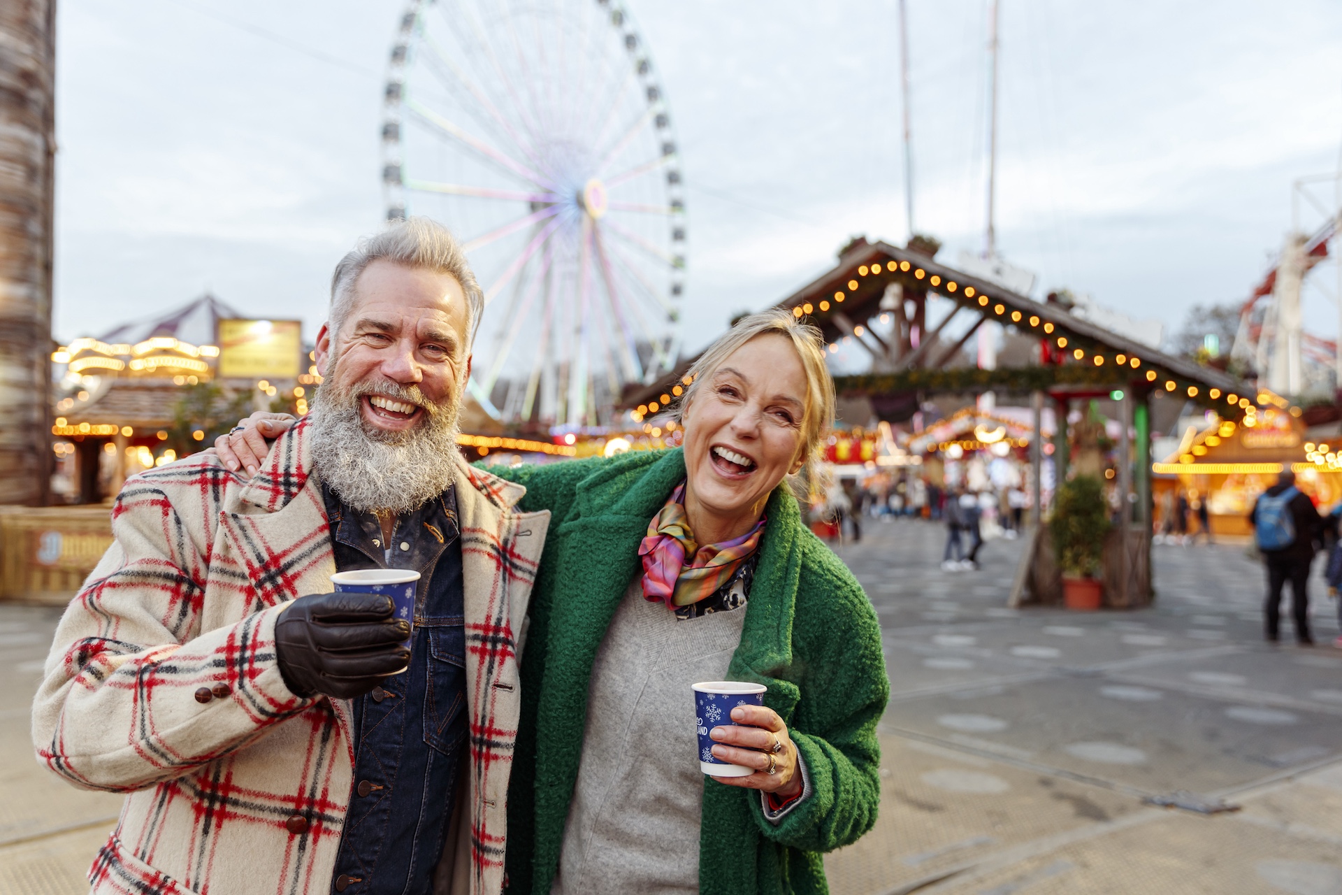 Paar mit warmen Getränken vor einem beleuchteten Riesenrad und Weihnachtsmarktbuden.