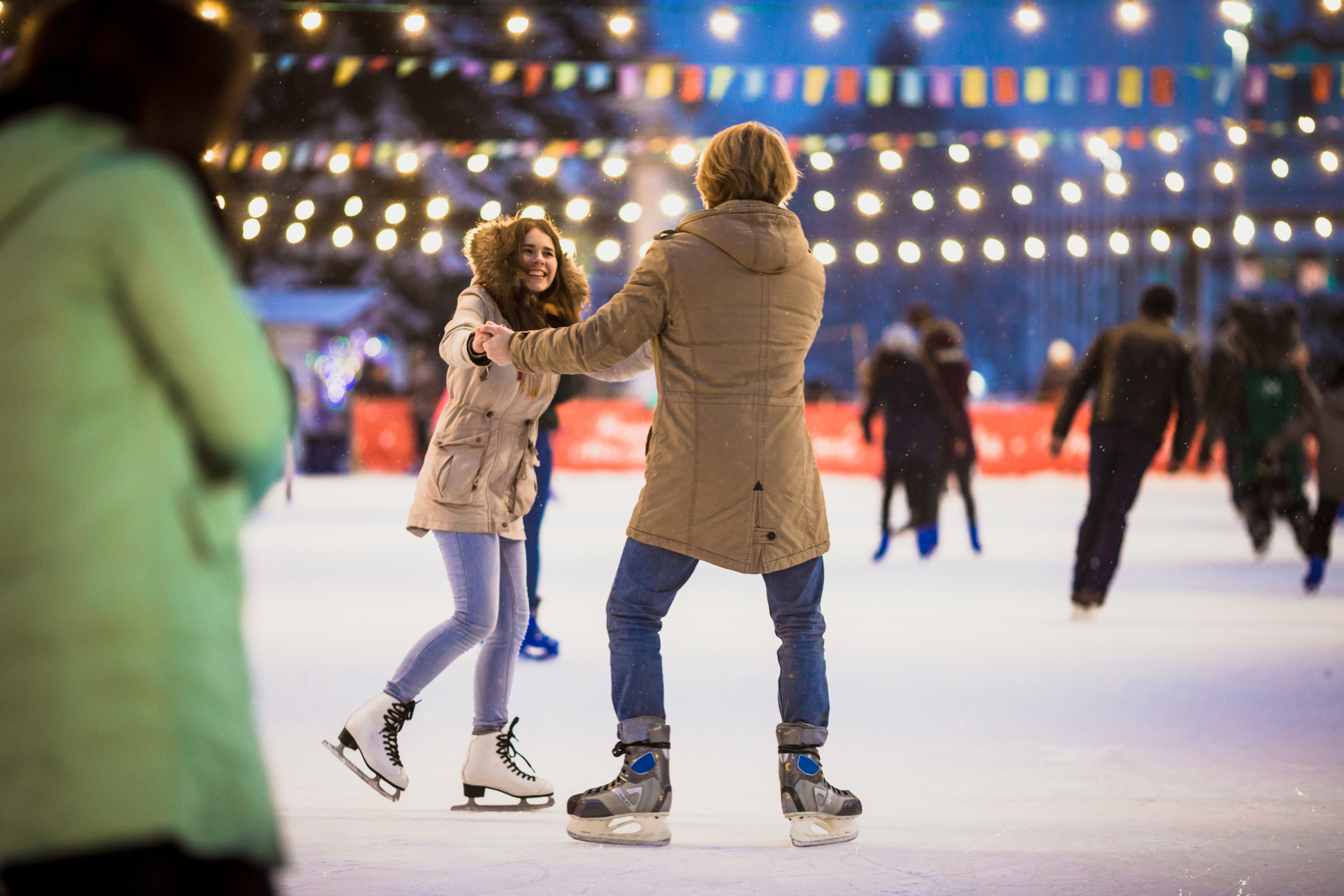 Zwei Personen beim Schlittschuhlaufen auf einer Eisbahn mit Lichterketten im Hintergrund.