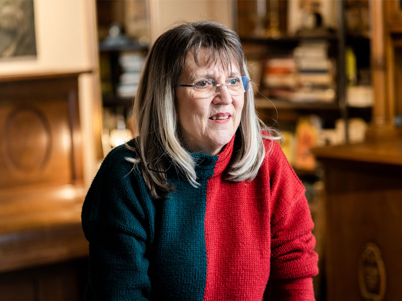 Eine Frau mit Brille in einem rot-grünen Pullover sitzt in einem gemütlichen Raum mit Bücherregal im Hintergrund.