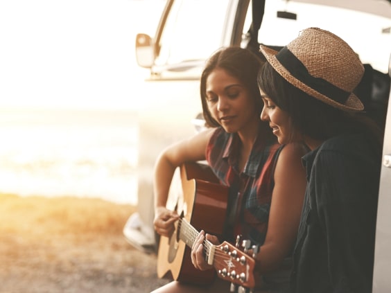 Zwei Frauen sitzen am offenen Van, eine spielt Gitarre, Sonnenuntergang im Hintergrund.