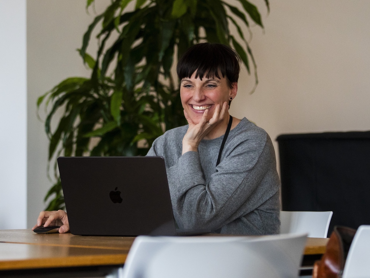 Frau mit grauem Pullover sitzt an einem Holztisch vor einem Laptop und hält die rechte Hand an das Kinn
