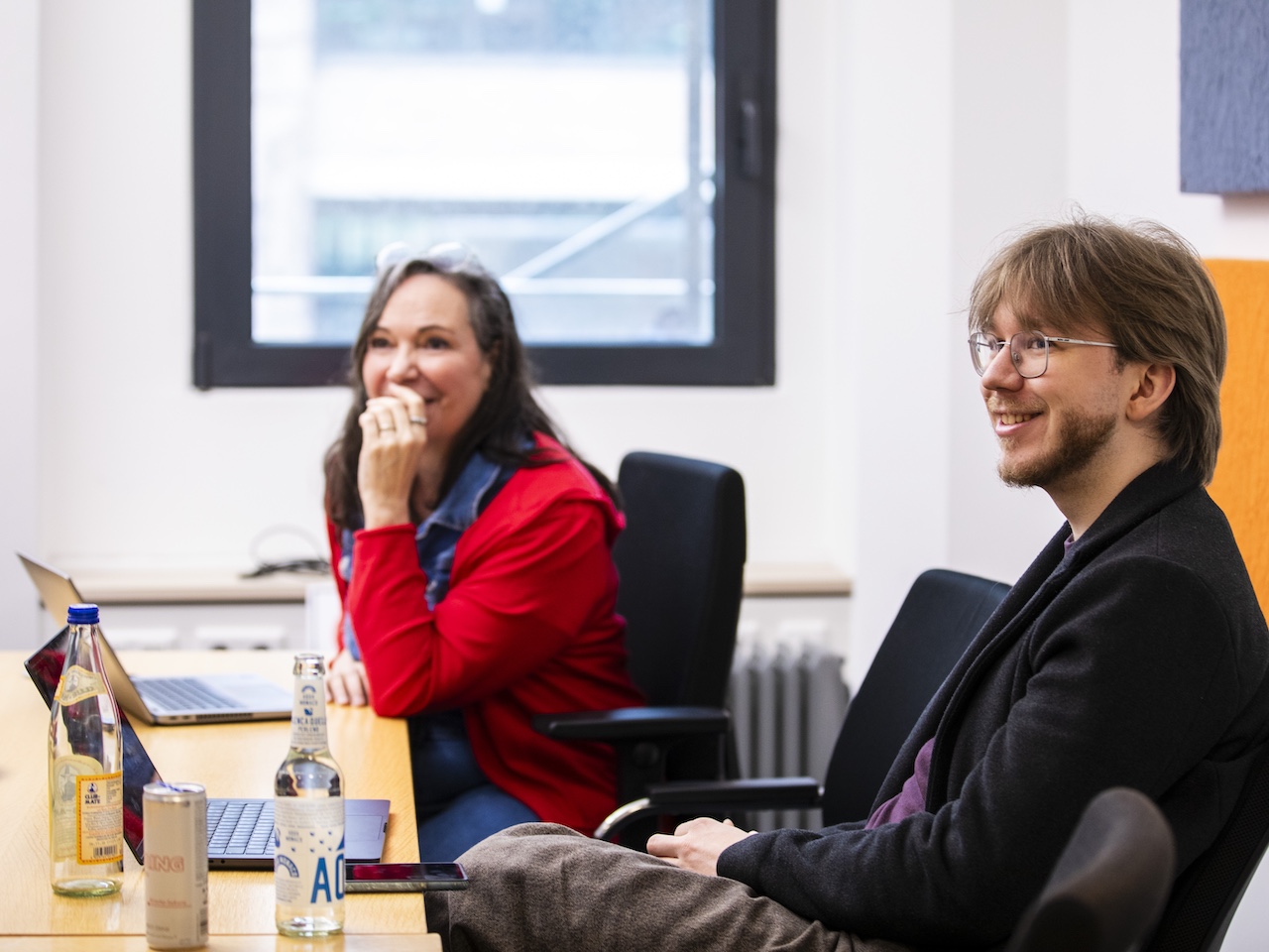 Zwei Personen sitzen in einem Büro an einem Tisch, eine trägt eine rote Jacke, die andere eine schwarze Jacke.