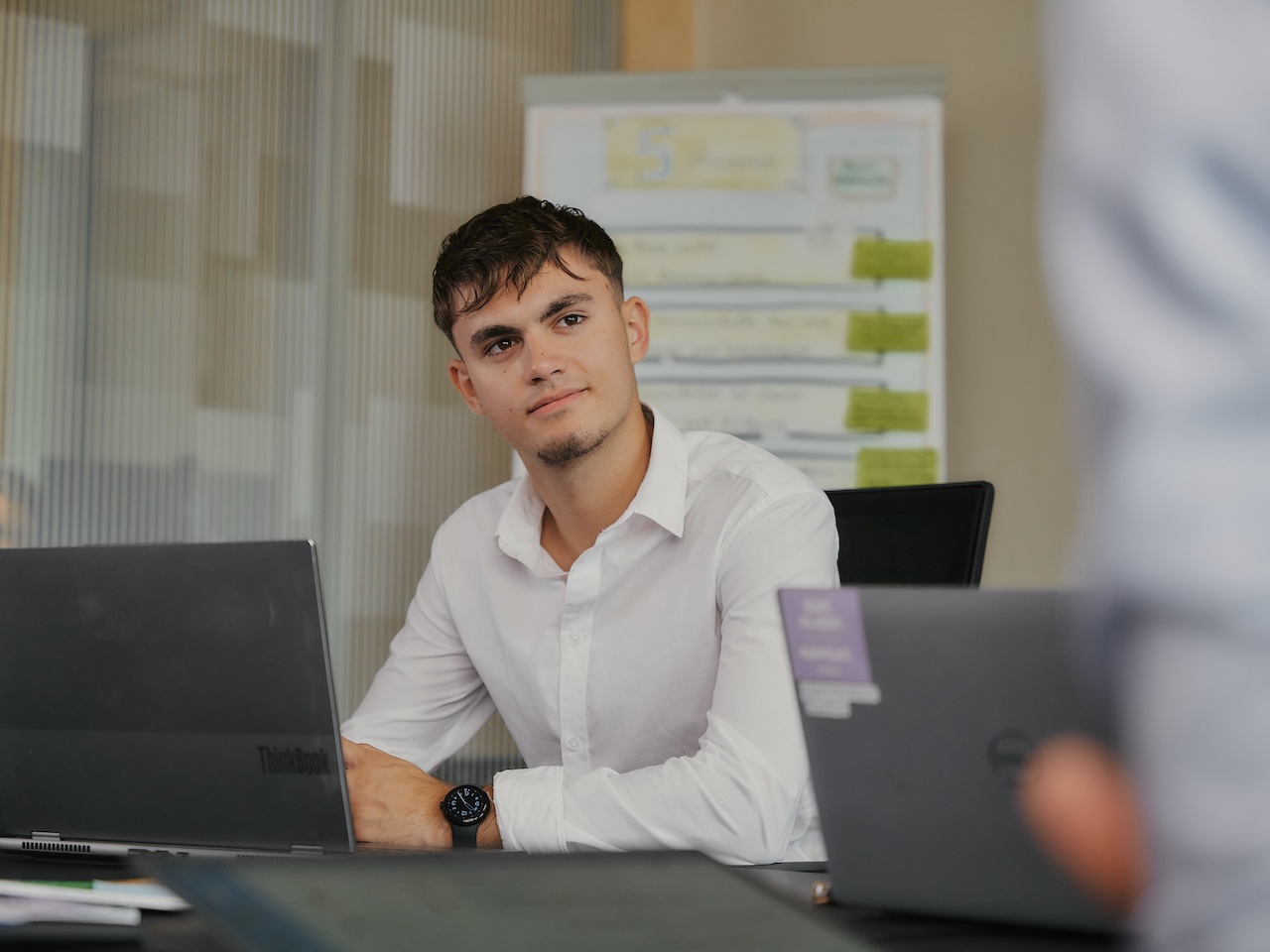Mann mit weißem Hemd sitzt an Schreibtisch mit zwei Laptops in Büro.