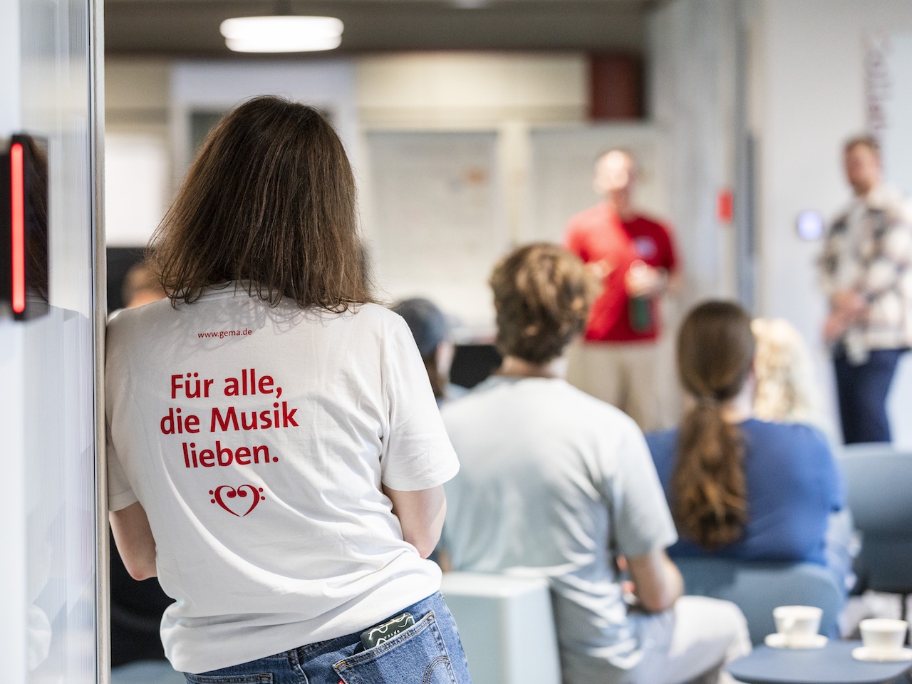 Rückenansicht einer Frau mit weißem T-Shirt und rotem Aufdruck 'Für alle, die Musik lieben.' in einem Raum mit mehreren sitzenden Personen.