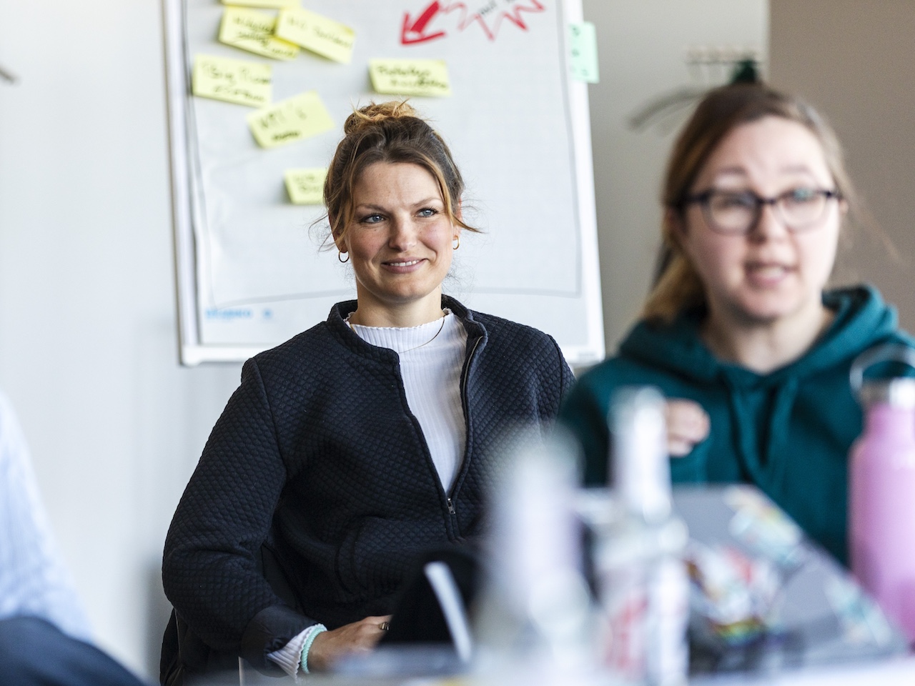 Zwei Frauen sitzen in einem Workshopraum vor einem Flipchart mit Notizzetteln, eine Frau trägt eine schwarze Jacke und lacht.