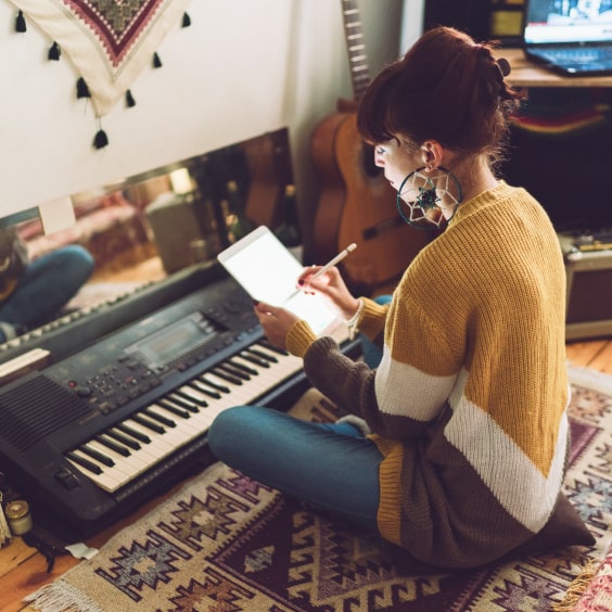 A woman is sitting on a carpet, writing on a tablet and has a keyboard in front of her.