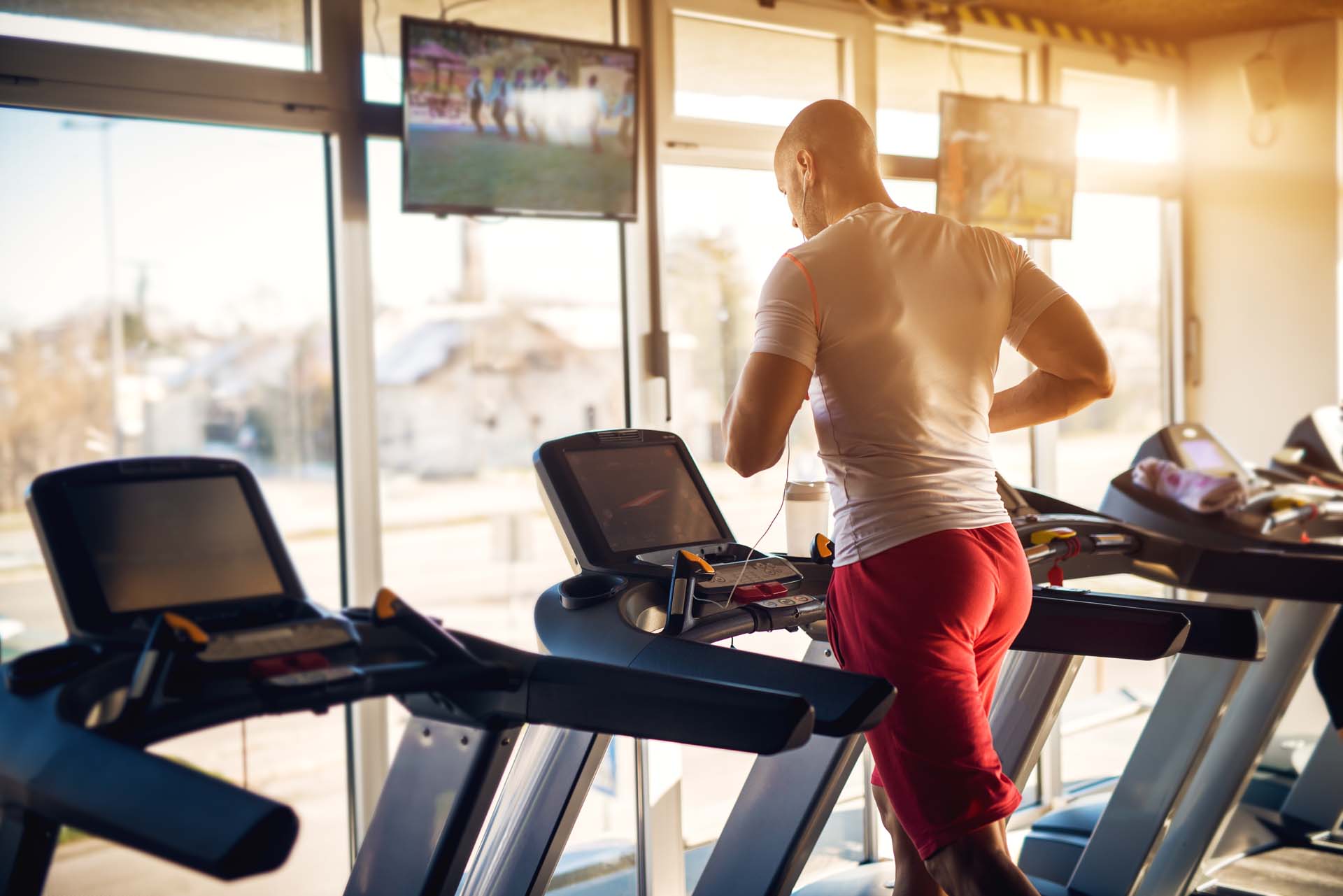 Ein Mann joggt auf einem Laufband im Fitnessstudio, mit Fenstern und Fernsehern im Hintergrund.