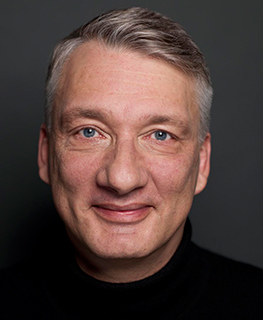 Eine lächelnde Person mit grauem Haar und schwarzem Rollkragenpullover vor dunkelgrauem Hintergrund.