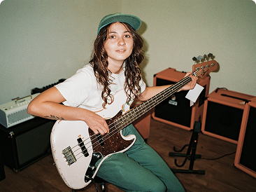 Eine junge Frau mit grüner Kappe spielt im sitzen Bass und blickt in die Kamera.