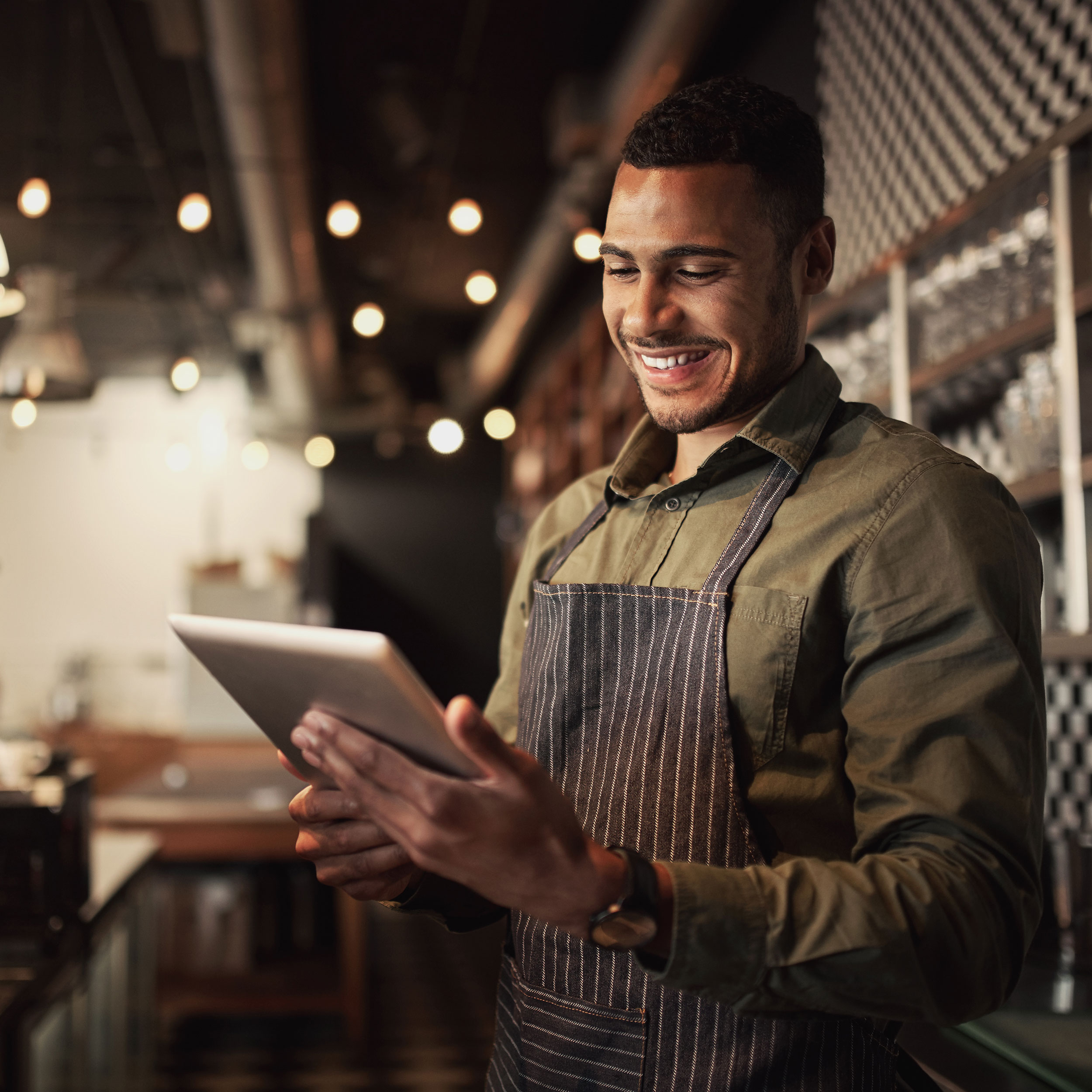 Ein Mann in Schürze lächelt und hält ein Tablet in einem gemütlichen Café mit warmem Licht.