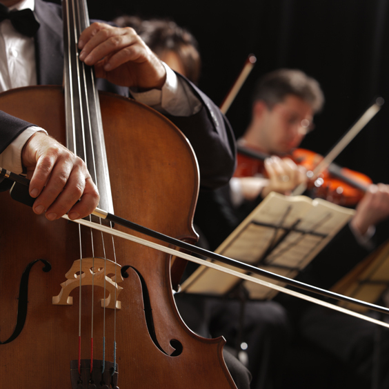 Ein Cellist spielt in einem Orchester, Notenständer und Geiger im Hintergrund.