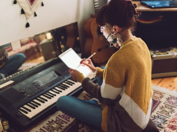 Eine Frau sitzt auf einem Teppich am Keyboard, schreibt auf einem Tablet, umgeben von Gitarren.