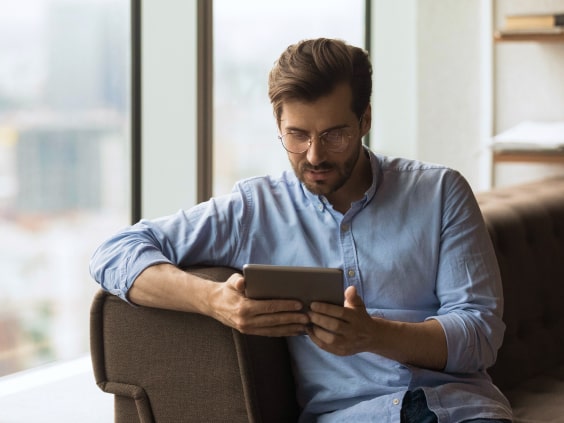 A man in a blue shirt sits on the sofa and reads on a tablet.